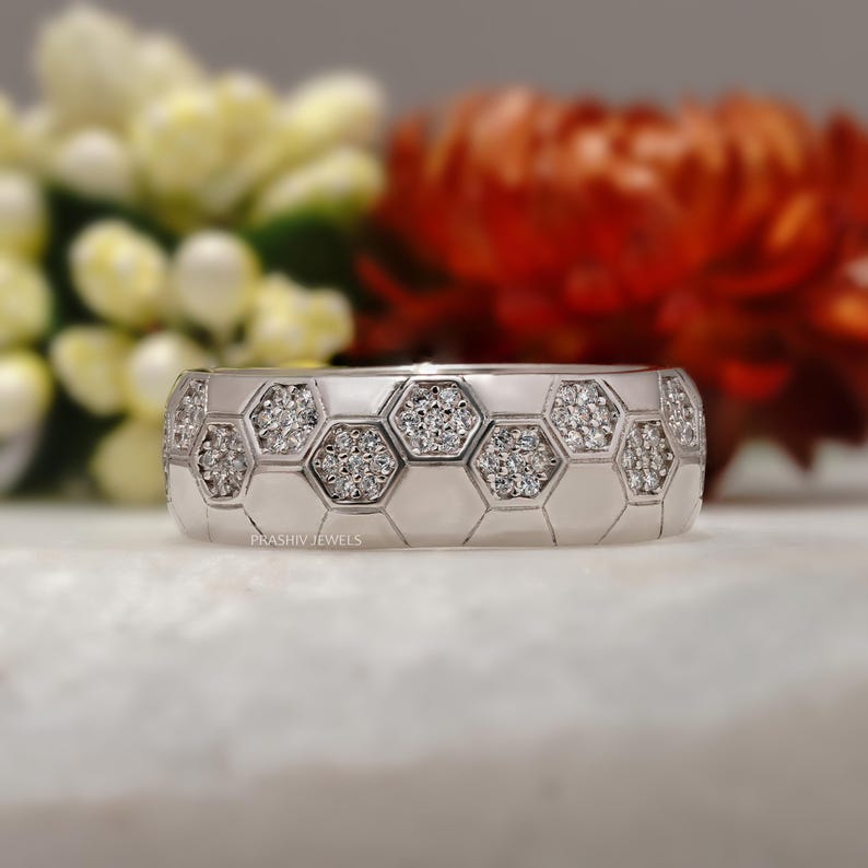 May include: A silver ring with a honeycomb design, each cell filled with small, sparkling diamonds. The ring is set against a blurred background of white and red flowers, highlighting the jewelry's intricate details and elegant style.