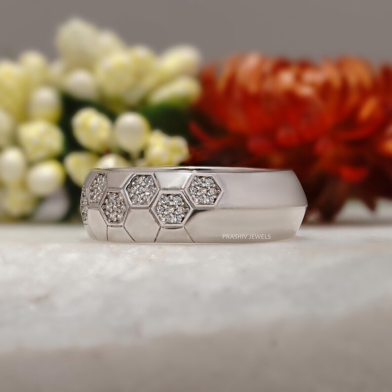 May include: A silver ring with a geometric honeycomb design. Each hexagon is filled with small, sparkling gemstones. The ring has a smooth, polished band and a modern aesthetic. The background features blurred flowers.