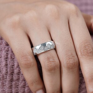 May include: A silver ring with a honeycomb design. Each hexagon is filled with small, sparkling stones. The ring is shown on a person's finger, with a soft pink background.