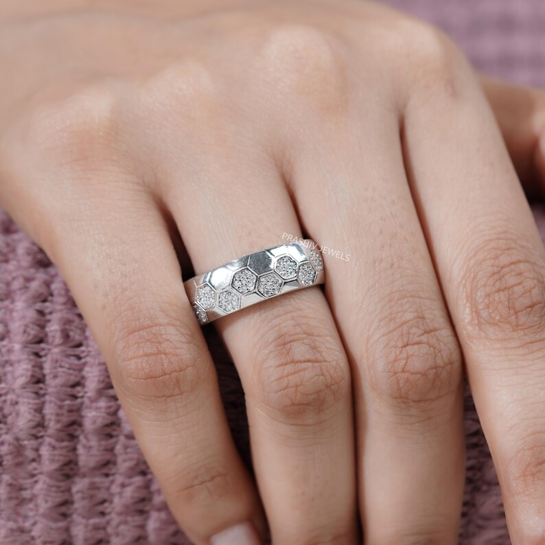 May include: A silver ring with a honeycomb design. Each hexagon is filled with small, sparkling stones. The ring is shown on a person's finger, with a soft pink background.