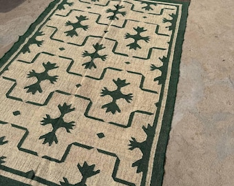 May include: A rectangular beige rug featuring a dark green geometric design and border. The rug has a fringe on the short ends. The pattern is made up of stylized floral shapes and angular lines.