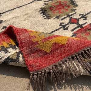 Puede incluir: Una alfombra tejida beige y roja con un patrón geométrico. La alfombra presenta una sección doblada con un diseño rojo, amarillo y marrón y flecos. El patrón incluye formas de diamantes negros y rojos.