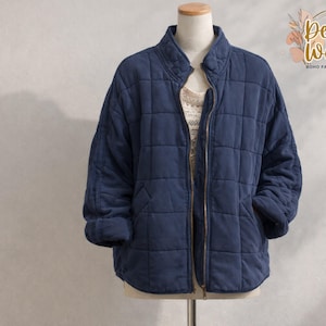 May include: A navy blue quilted jacket with a stand-up collar and a full-length zipper. The jacket has a square quilted pattern and two front pockets. The jacket is displayed on a mannequin. The Desert Weave Boho Fashion Boutique logo is visible in the upper right corner.