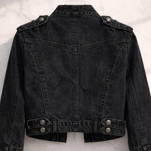 May include: A black denim jacket with a classic biker style. The jacket features a notched collar, shoulder epaulets, and silver-tone button details. The back of the jacket has a tailored fit with seams and a belt detail at the waist.