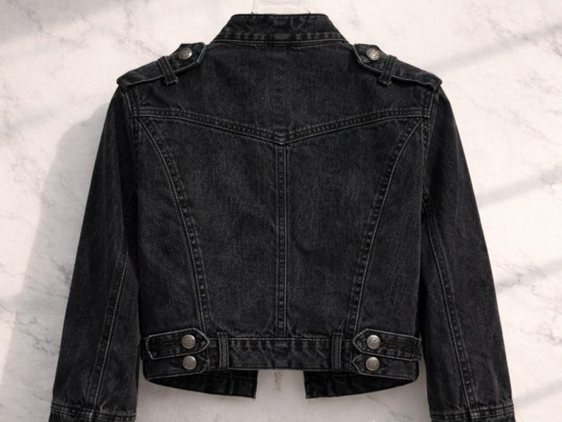 May include: A black denim jacket with a classic biker style. The jacket features a notched collar, shoulder epaulets, and silver-tone button details. The back of the jacket has a tailored fit with seams and a belt detail at the waist.