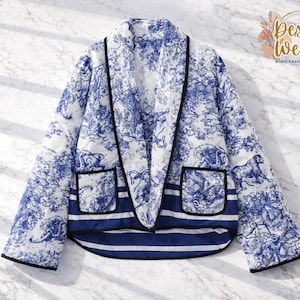 May include: A quilted jacket in white and blue, featuring a toile de Jouy pattern with horses and pastoral scenes. The jacket has black trim, two pockets, and a striped hem. The Desert Weave Boho Fashion Boutique logo is visible.