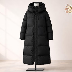 May include: A long, black puffer coat with a hood, displayed on a mannequin. The coat is floor-length and has a quilted design. The coat has a zip closure and button details. The background is a neutral colour.