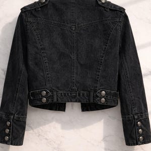 May include: A black denim jacket with silver-toned buttons. The jacket features a classic design with a tailored fit, including buttoned cuffs and a structured collar. The back of the jacket has a detailed yoke and paneling.