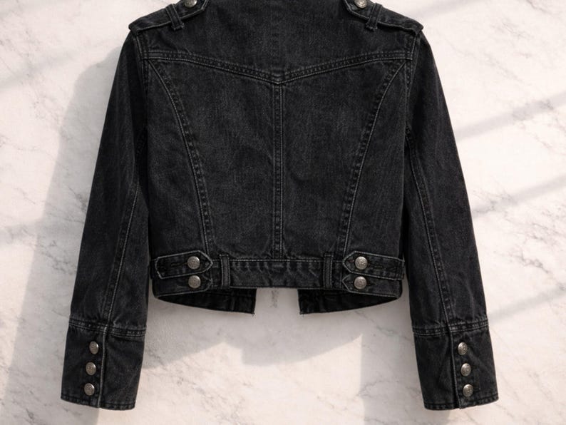 May include: A black denim jacket with silver-toned buttons. The jacket features a classic design with a tailored fit, including buttoned cuffs and a structured collar. The back of the jacket has a detailed yoke and paneling.