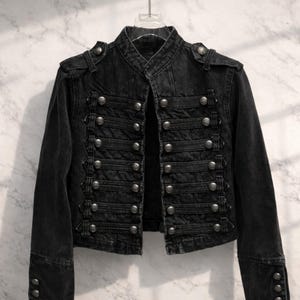 May include: Black denim jacket with a military-inspired design. The jacket features a mandarin collar, multiple rows of silver-colored buttons, and epaulettes on the shoulders. The sleeves have buttoned cuffs. The jacket is hanging against a marble-patterned background.
