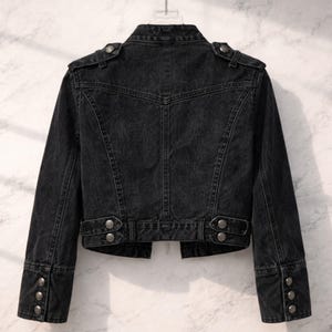 May include: Back view of a black denim jacket. The jacket features a classic design with buttoned cuffs, shoulder epaulettes, and decorative buttons on the waist. The jacket is hanging on a hanger against a light background.