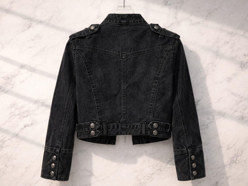 May include: Back view of a black denim jacket. The jacket features a classic design with buttoned cuffs, shoulder epaulettes, and decorative buttons on the waist. The jacket is hanging on a hanger against a light background.