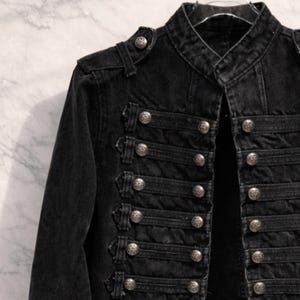 May include: Black denim jacket with a military-inspired design. The jacket features a mandarin collar, epaulets, and multiple rows of decorative silver buttons. The jacket has long sleeves and a tailored fit, suitable for various occasions.