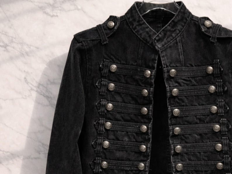 May include: Black denim jacket with a military-inspired design. The jacket features a mandarin collar, epaulets, and multiple rows of decorative silver buttons. The jacket has long sleeves and a tailored fit, suitable for various occasions.