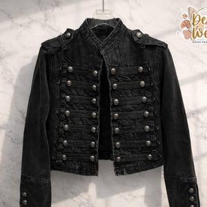 Puede incluir: Chaqueta vaquera negra con múltiples filas de botones plateados y cuello mandarín. La chaqueta tiene charreteras en los hombros y puños abotonados. El logotipo de Desert Weave Boho Fashion Boutique es visible.