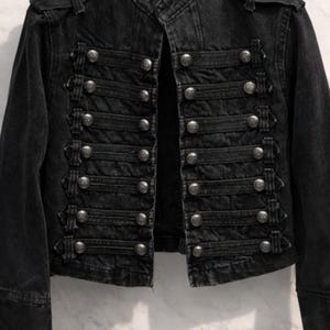 May include: Black denim jacket with a military-inspired design. The jacket features multiple rows of silver-colored buttons and decorative straps. The jacket has a structured fit with a high collar and long sleeves.