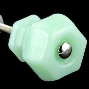 Pode incluir: Um puxador de móvel hexagonal em vidro verde menta pálido com acabamento liso e brilhante. O centro do puxador apresenta um pequeno detalhe metálico circular em tom prateado polido. Um parafuso roscado e uma porca quadrada estão fixados atrás.