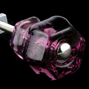 Pode incluir: Um botão de armário de vidro roxo com um parafuso de metal prateado. O botão tem um design facetado e uma base redonda de metal prateado.