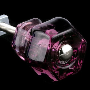 Puede incluir: Un pomo de armario de vidrio morado con un tornillo de metal plateado. El pomo tiene un diseño facetado y una base redonda de metal plateado.