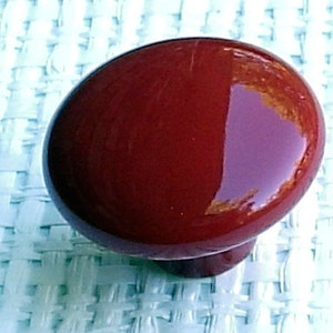 May include: A glossy, dark red cabinet knob with a rounded top and a smaller, cylindrical base. The knob reflects light, showing a glimpse of the surroundings. This hardware piece is likely for furniture or cabinetry.