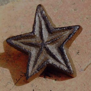 May include: A cast iron star with a rustic, weathered appearance. The five-pointed star has a dark brown color with lighter brown highlights, and a textured surface. The star is likely a decorative item or a piece of hardware.