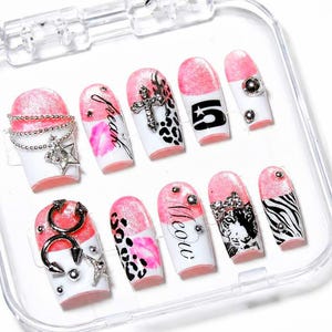 May include: A collection of ten false nails presented in a clear case. The nails have a white base with pink tips and diverse designs, such as leopard print, zebra stripes, and the word "meow". Some nails are adorned with silver embellishments.