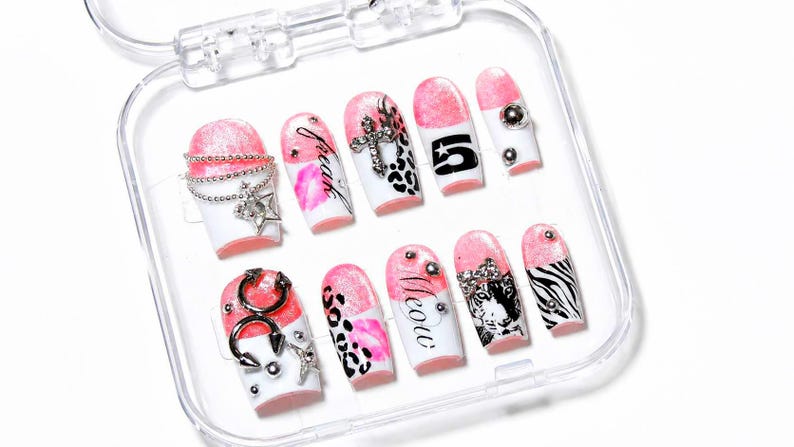 May include: A collection of ten false nails presented in a clear case. The nails have a white base with pink tips and diverse designs, such as leopard print, zebra stripes, and the word "meow". Some nails are adorned with silver embellishments.