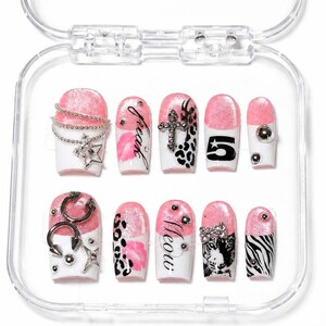 May include: A collection of ten false nails presented in a clear case. The nails showcase a pink and white design, incorporating elements such as silver chains, stars, crosses, leopard print, the number 5, and the words "freak" and "meow".