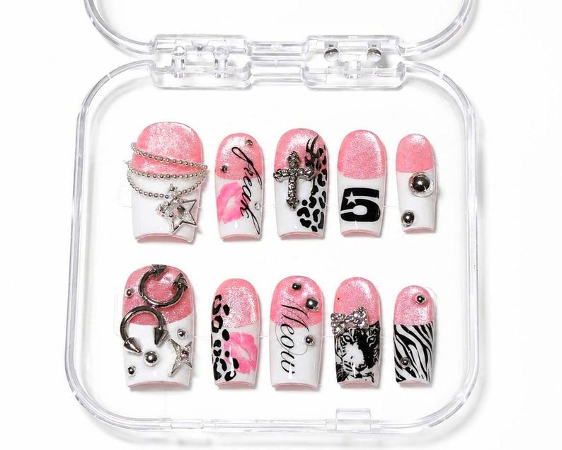 May include: A collection of ten false nails presented in a clear case. The nails showcase a pink and white design, incorporating elements such as silver chains, stars, crosses, leopard print, the number 5, and the words "freak" and "meow".