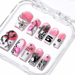 May include: A set of false nails in a clear plastic case. The nails have a white base with pink tips and various designs, including silver charms, leopard print, and the word "freak".