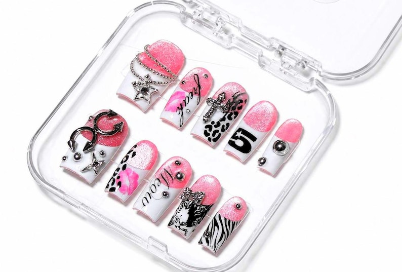 May include: A set of false nails in a clear plastic case. The nails have a white base with pink tips and various designs, including silver charms, leopard print, and the word "freak".