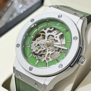 以下が含まれることがあります： 緑色の文字盤と緑色の革ストラップが付いた、シルバーカラーのHublotの腕時計。文字盤は透明で、内部のメカニズムが見えます。白い箱に入っています。