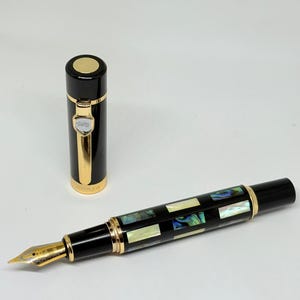 Könnte beinhalten: Ein schwarzer Füllfederhalter mit goldenen Akzenten und einem dekorativen Abalone-Inlay. Die Kappe ist abgenommen und zeigt eine goldene Feder. Es ist ein luxuriöses Schreibgerät.