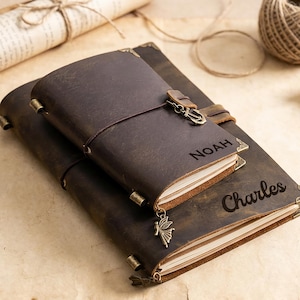 May include: Two brown leather journals, one stacked on top of the other. The top journal is smaller and has the name "Noah" engraved on it. The bottom journal has the name "Charles" engraved. Both have metal corner protectors and clasps.