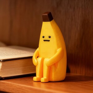 Könnte beinhalten: Gelbe, bananenförmige Figur mit brauner Spitze und einfachem Gesicht. Die Figur sitzt auf einem Holzregal neben einem offenen Buch. Die Banane hat Arme und Beine und sitzt.