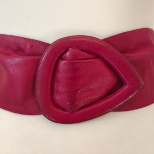 Op de afbeelding: Een brede, fuchsia leren riem met een grote, afgeronde gesp. De riem wordt getoond op een mannequin, waarbij de vorm en het ontwerp worden benadrukt. De gesp is een belangrijk kenmerk en voegt een gedurfd accent toe aan het accessoire. De riem is ongeveer 80 cm lang.