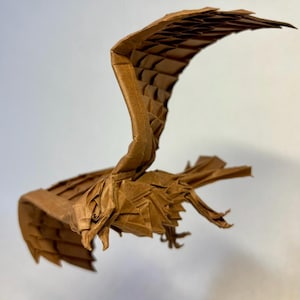 Puede incluir: Un pájaro de origami marrón en vuelo, con detalles de pliegues de alas y plumas. La escultura de papel se captura en el aire sobre un fondo liso, mostrando la intrincada artesanía del papel plegado.