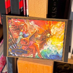 May include: A colorful fantasy art print featuring a warrior figure riding a dragon, displayed on a wooden easel. The artwork is set against a backdrop of bookshelves filled with books, including titles like "Monster Manual" and "The Art of Cuphead".