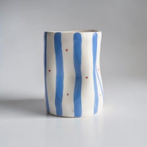 Könnte beinhalten: Eine weiße Keramikvase mit vertikalen blauen Streifen und kleinen roten Herzakzenten. Die Vase hat eine leicht unregelmäßige Form und eignet sich zum Halten von Blumen oder als dekoratives Stück.