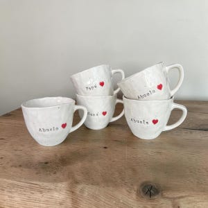 Może przedstawiać: Cztery białe kubki ceramiczne o nakrapianej fakturze. Każdy kubek posiada małe czerwone serce oraz czarny napis. Na kubkach widnieją słowa Abuelo, Papa, Mama oraz Abuela, a każdemu z nich towarzyszy czerwona ikona serca na przedniej powierzchni.