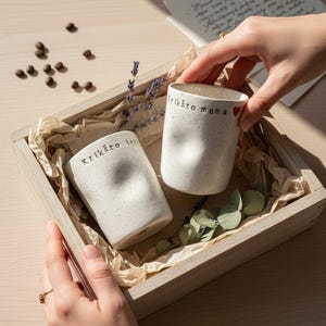 Peut inclure: Deux tasses en céramique blanc cassé avec des textures mouchetées et des côtés indentés, nichées dans une boîte en bois avec du papier froissé. Une tasse porte le texte Krikšto mama et un cœur rouge, l'autre le texte Krikšto tėt.