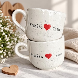 Puede incluir: Dos tazas de cerámica blanca con motas negras, cada una con un asa curva. La taza superior muestra el texto negro Krikšto y Tėtis con un corazón rojo en medio. La taza inferior muestra el texto negro Krikšto y Mama con un corazón rojo.