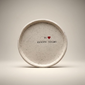 May include: A round, speckled, off-white ceramic tray with a raised edge. The tray features the text "SU" above a red heart, and the text "KRIKŠTO TEVAMS" below the heart. The tray has a minimalist design.