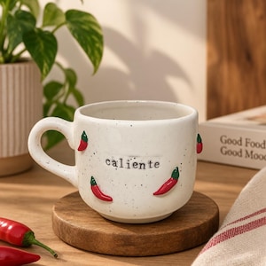 Puede incluir: Una taza de cerámica blanca con acabado moteado, decorada con pequeños chiles rojos en relieve y la palabra "caliente" impresa en letras minúsculas negras en el frente. La taza cuenta con un asa redondeada y una base circular ancha.