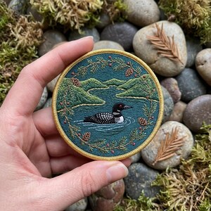 May include: Round embroidered patch featuring a loon swimming in a lake, surrounded by green hills and pine branches. The patch has a teal background and a gold border. The loon is black and white with a red eye. The patch is held in a hand.