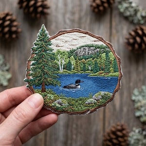 May include: Embroidered patch featuring a serene lake scene with a loon, trees, and a mountain. The patch is framed by a brown border resembling a tree cross-section. The patch is held in a hand, with pine cones in the background.