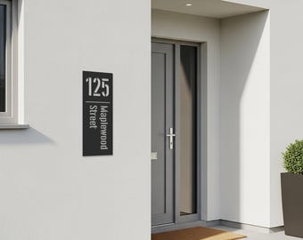 May include: A black metal address sign with the number "125" at the top and "Maplewood Street" written vertically below. The sign is mounted on a white wall next to a gray door.