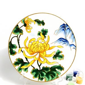 Può includere: Un piatto decorativo in ceramica bianca con un vivace motivo floreale di crisantemo giallo, foglie verdi e dettagli di montagne blu. Il piatto ha un bordo dorato ed è esposto su un supporto trasparente. Sono visibili piccoli vasetti di vernice.