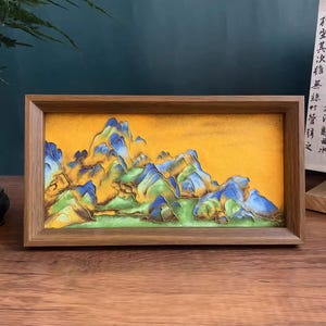 May include: A framed landscape painting featuring a mountain range with blue, green, and yellow hues. The artwork is set against a yellow background and enclosed in a wooden frame, creating a decorative piece.