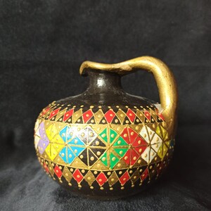 Op de afbeelding: Een kleine, ronde, zwarte keramische vaas met een gouden handvat. De vaas is versierd met een geometrisch patroon van rode, gele, groene, blauwe en witte ruiten en driehoeken, omlijnd met goud. De vaas heeft een kleine opening aan de bovenkant.